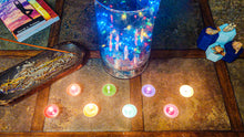 Load image into Gallery viewer, Chakra healing tealight candles lit and displayed on a table along with incense, books and other meditation and altar space items.